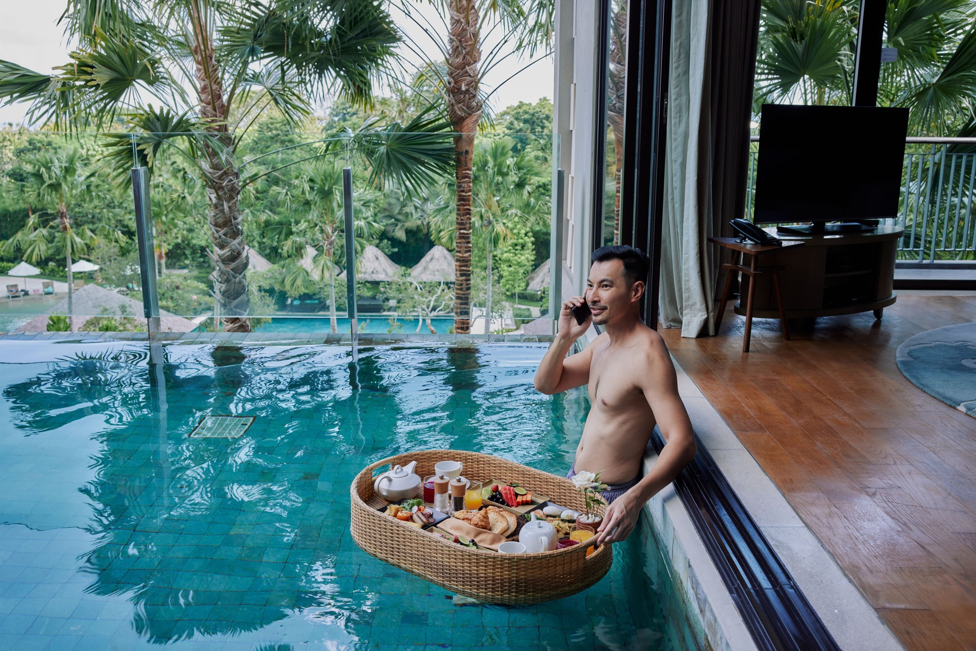 Happy man in swimsuit having floating breakfast tray in luxury pool hotel while talking on the phone. Young male enjoying morning coffee in tropical resort and working remotely on online project